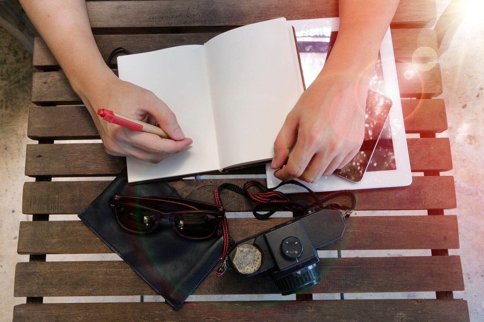 Overhead view of hands writing in an open blank notebook beside a camera and sunglasses